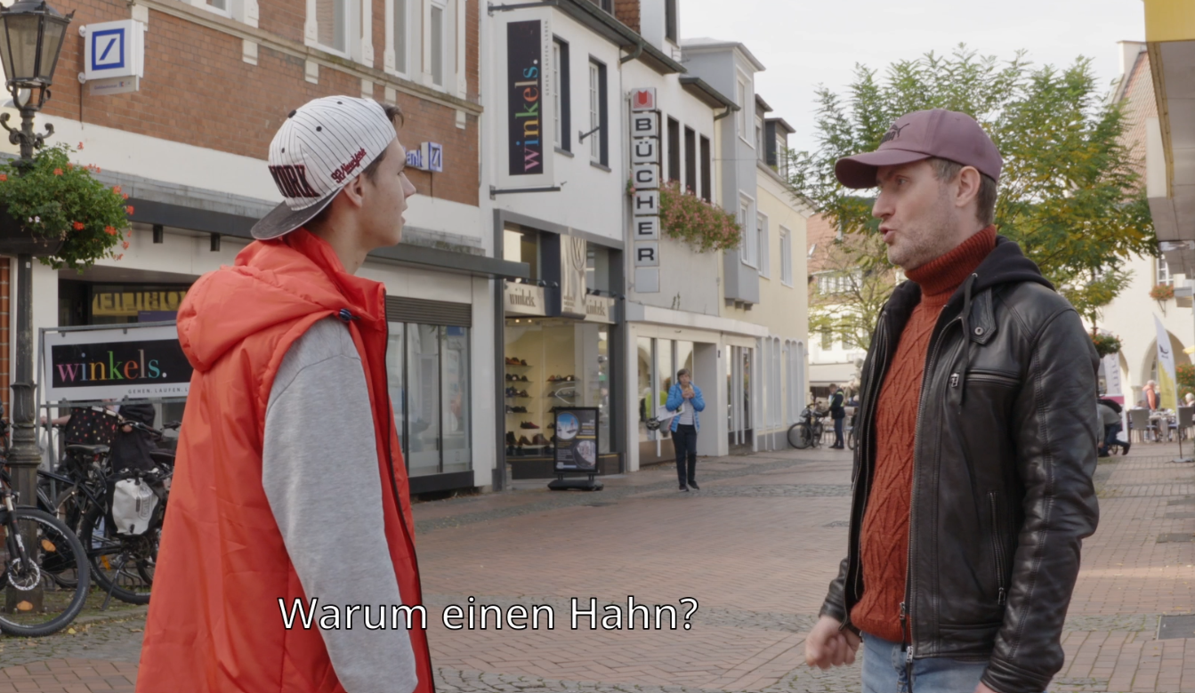 Interviewsituation: Zwei junge Männer in einer Innenstadt. Untertitel "Warum einen Hahn?"